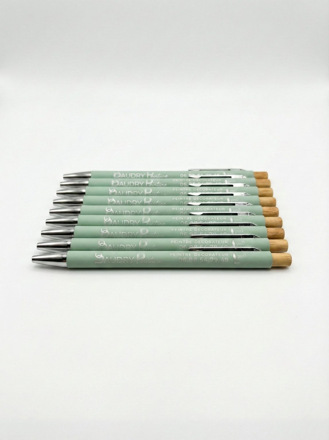 Alignement de stylos vert pastel personnalisés “Baudry Peinture” avec embout bois, présentés sur fond blanc.