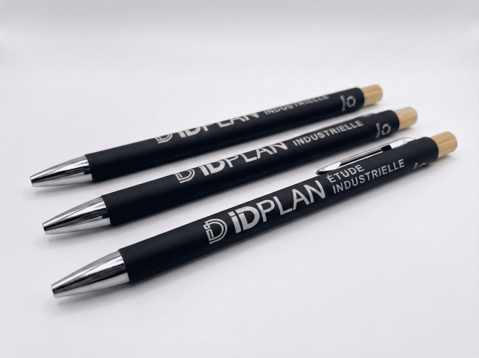 Trois stylos noirs personnalisés “IDPLAN – Étude industrielle” avec embout bois, présentés sur fond blanc.